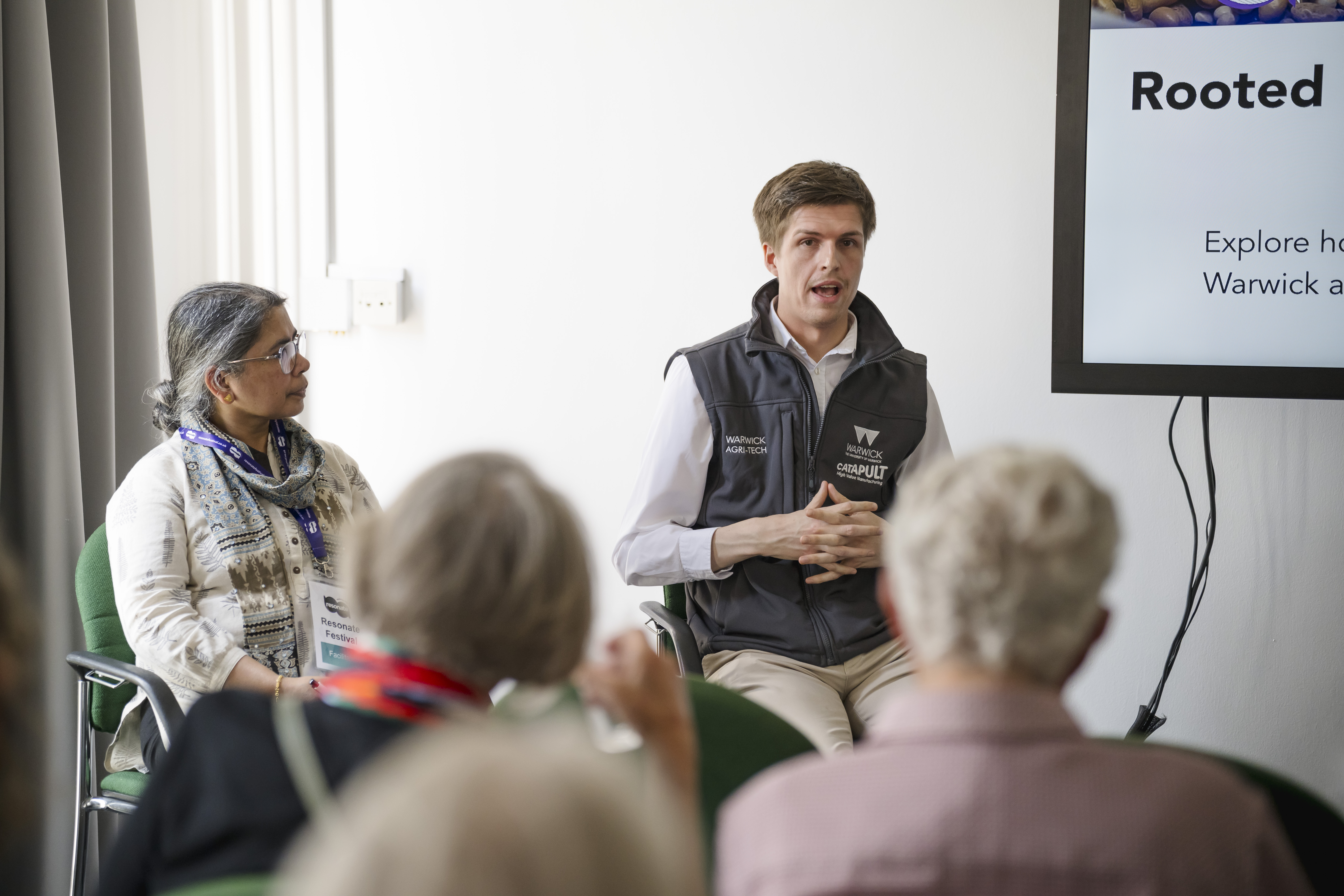 A man from the Agritech team at the University of Warwick is sat at the front of a room, speaking to an audience. Next to him is a woman from Feeding Coventry who is watching him speak.
