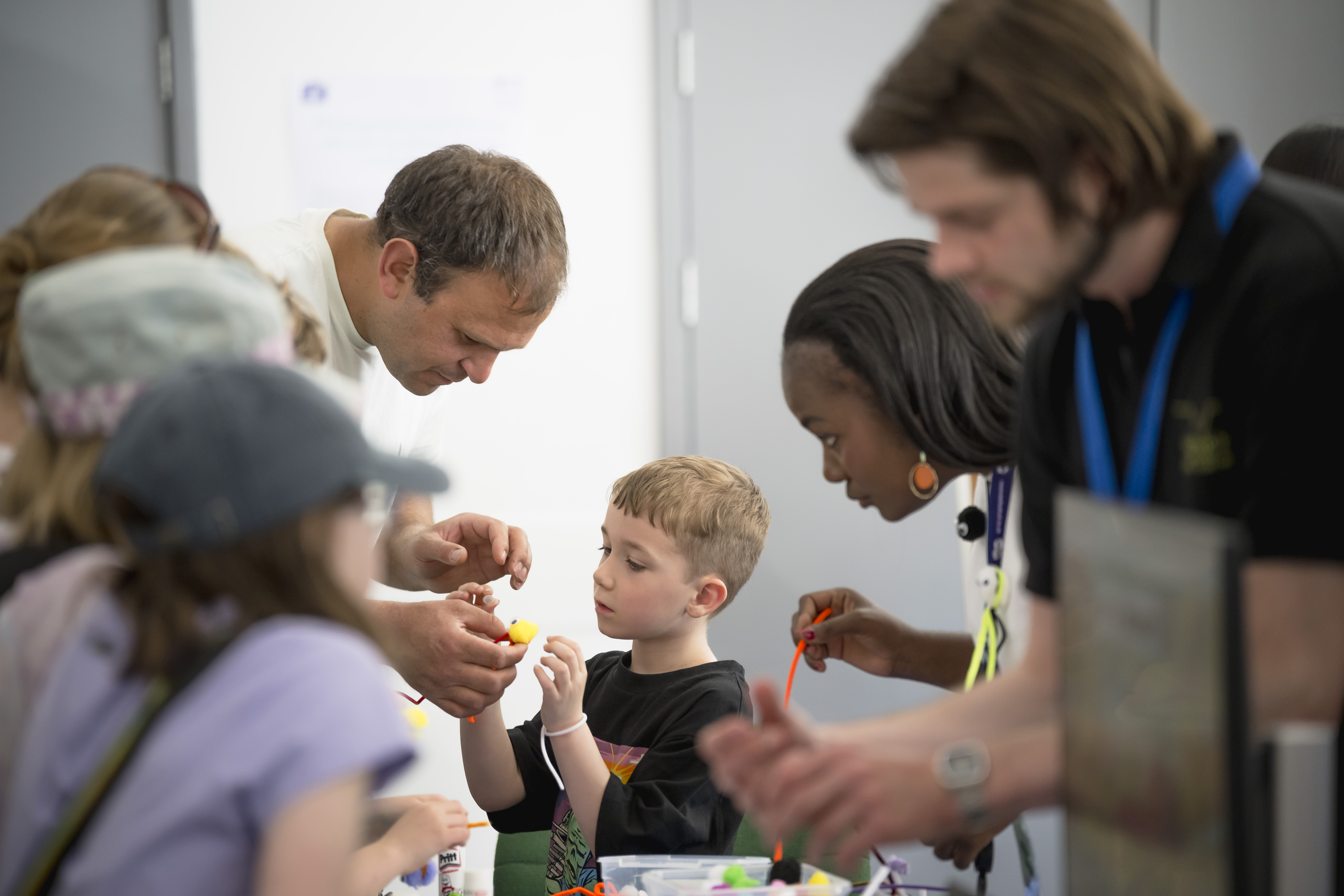 A group of people are gathered around a table, making plant pathogens out of pipe cleaners. The image is focused on an adult and a child making their craft.