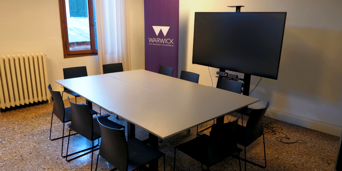 A seminar room with 12 chairs around a table, in front of a large digital screen and camera setup