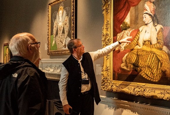 A volunteer points to a painting as visitors look on