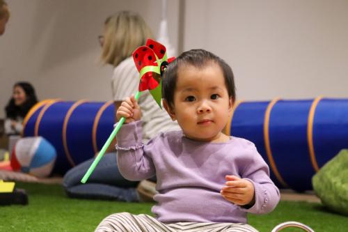 A small child holds a windmill toy whilst sat in a play area