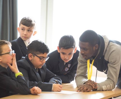 A student volunteer leans on a desk to talk to four pupils as aprt of the raising aspiration project
