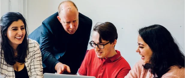 A member of staff is pointing to a laptop screen, explaining something to three students who are all smiling