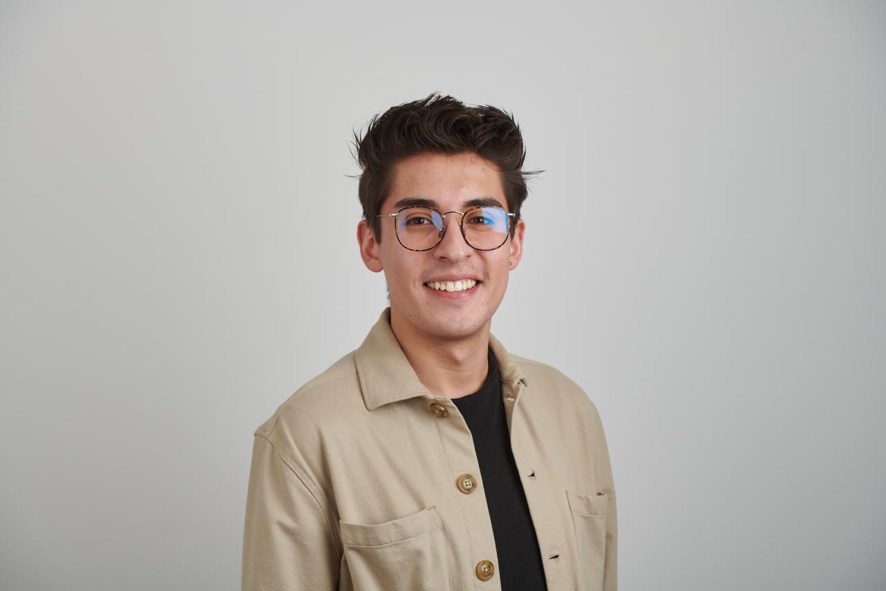 Daniel smiles at camera wearing glasses and a beige jacket