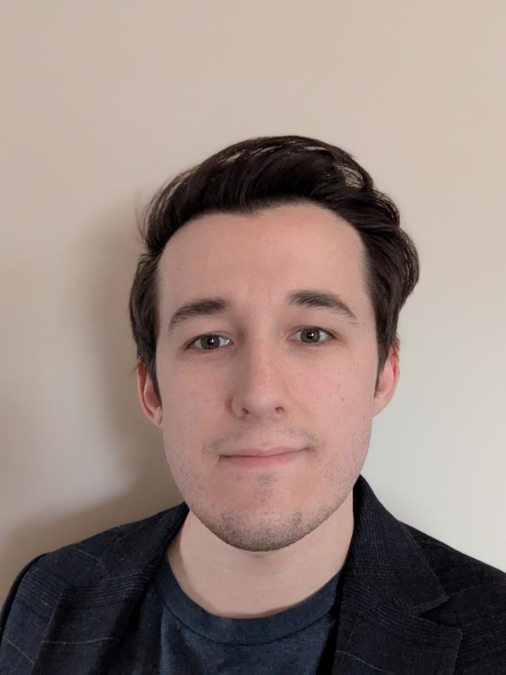 Dylan stands in front of a beige wall with a black blazer and grey t-shirt