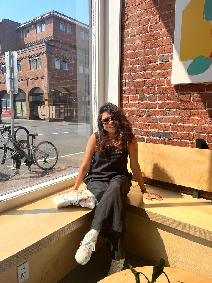 Elvira sits in a cafe window with the sun on her. She smiles in sunglasses