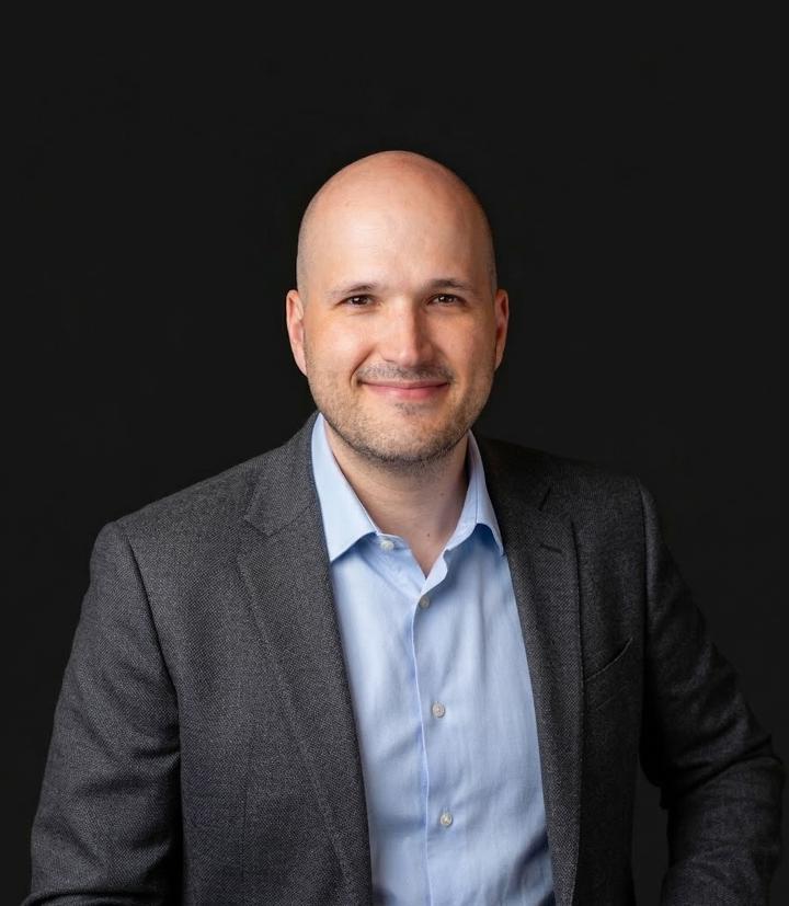Julien looks directly at camera with a small smile. He wears a mid-grey blazer and light blue shirt