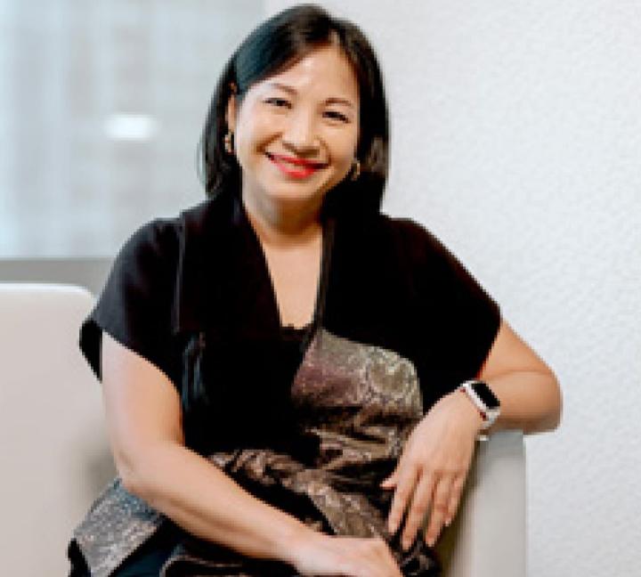 Karin sits on a off-white sofa wearing a black dress with a decorative bow. She smiles at camera