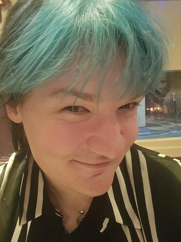 Lizzie has blue hair and smiles at the camera wearing a black and white striped shirt