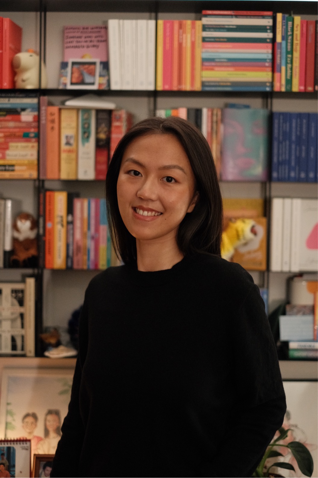 Ming stands in a black jumper before a bookcase filled with colourful books