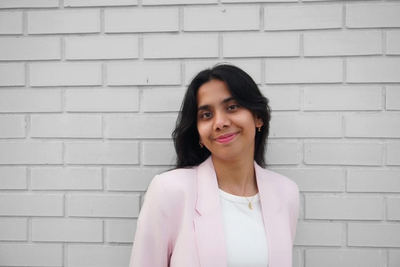 Saumya stands beside a white brick wall wearing a pale pink blazer