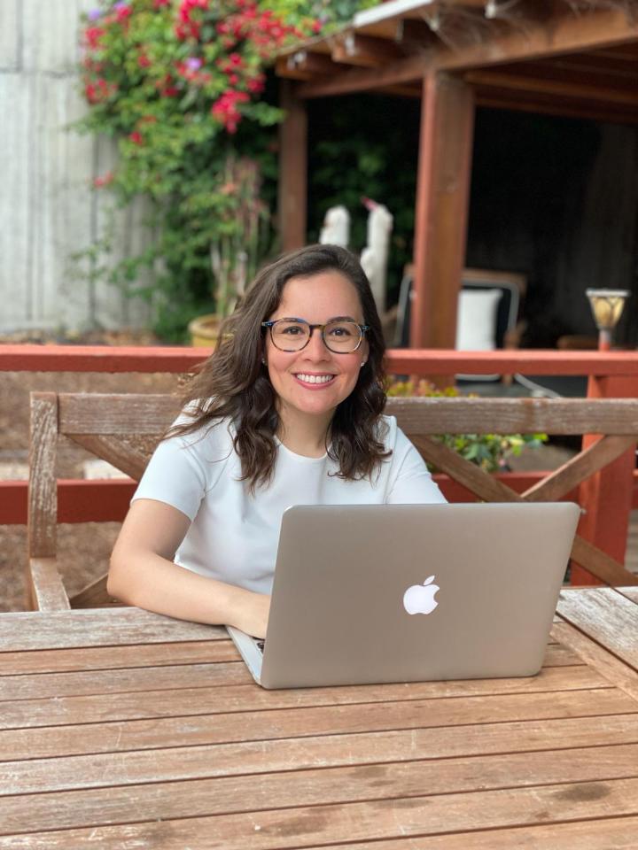 Sebnem sits outside with her Macbook open and a pair of glasses on her face smiling