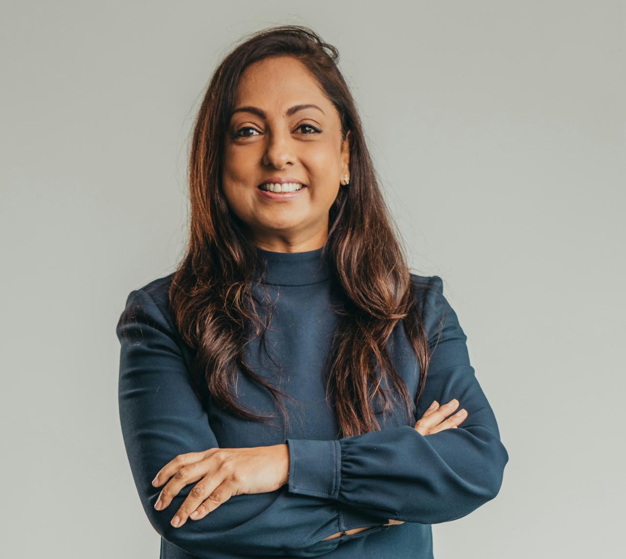 Vandana wears a deep blue jumper and stands with her arms crossed