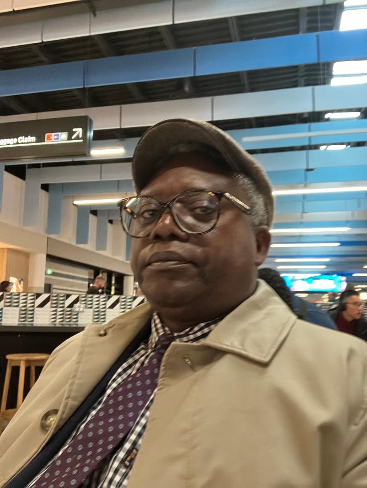 Charles at an airport in a flat cap and spotty tie