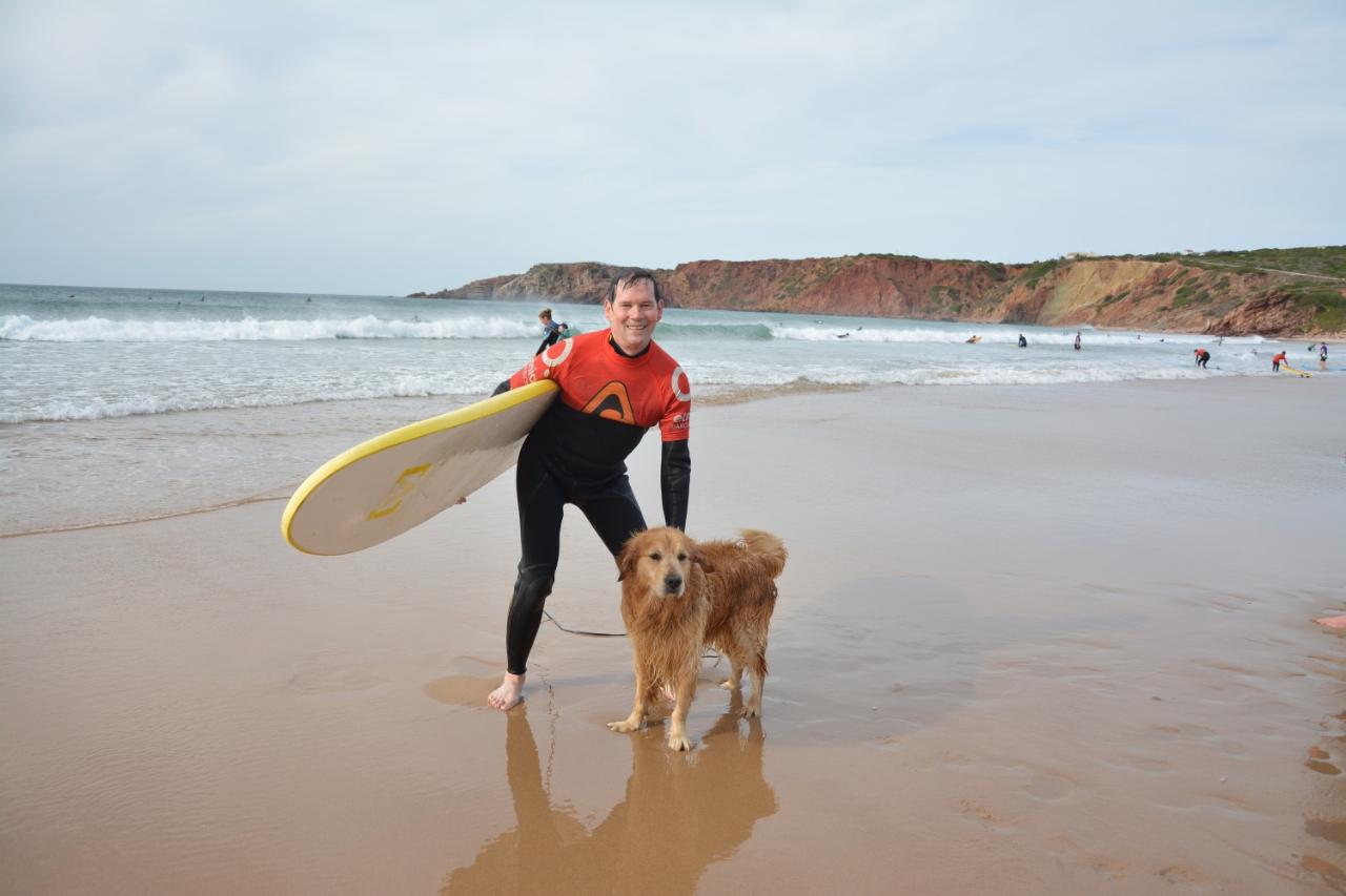 Andrew Spence stands on a beach holding a surfboard and reaching down to a dog