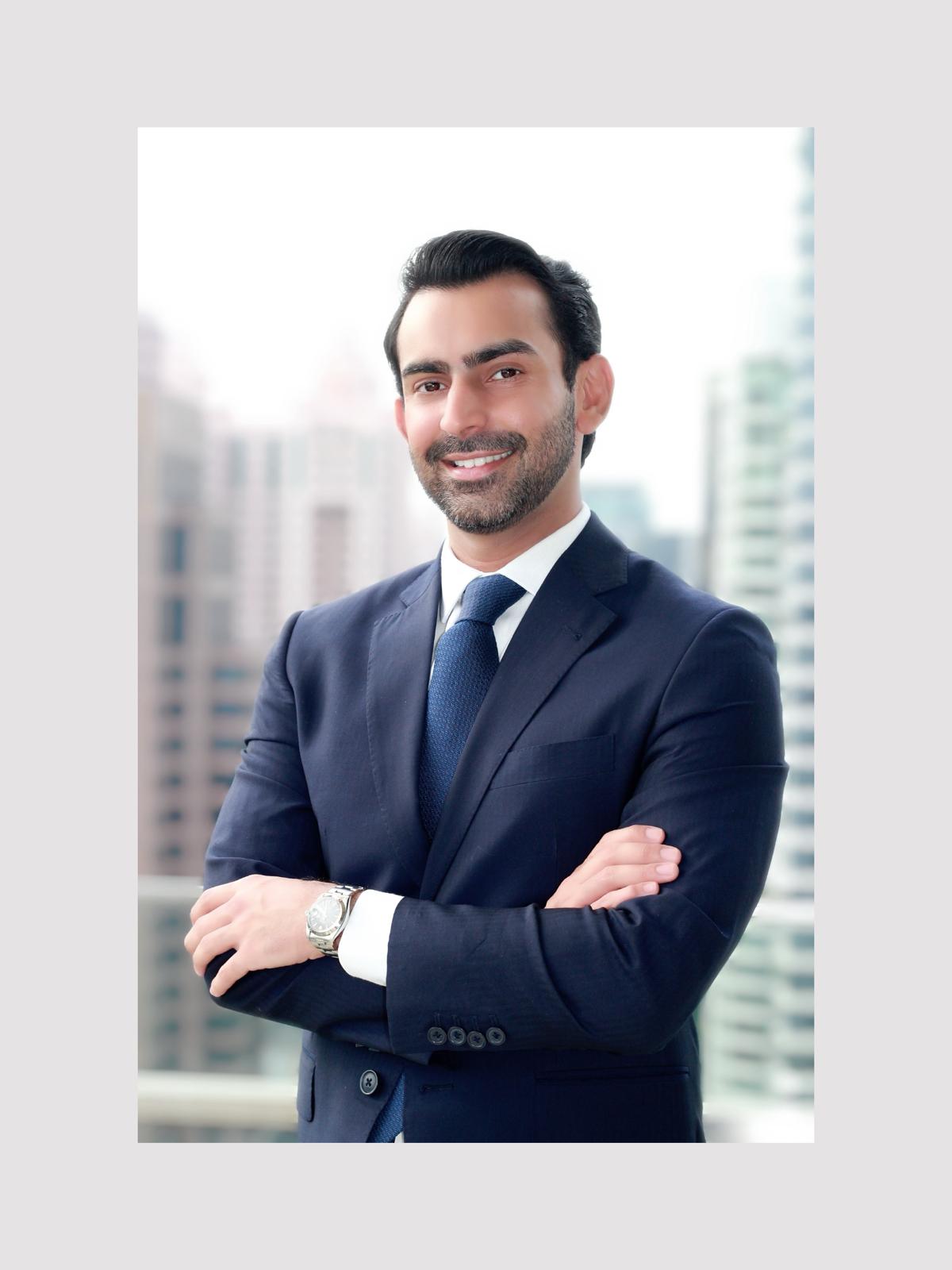 Abbas wears a navy suit and blue tie. He has his arms crossed and stands in front of a cityscape