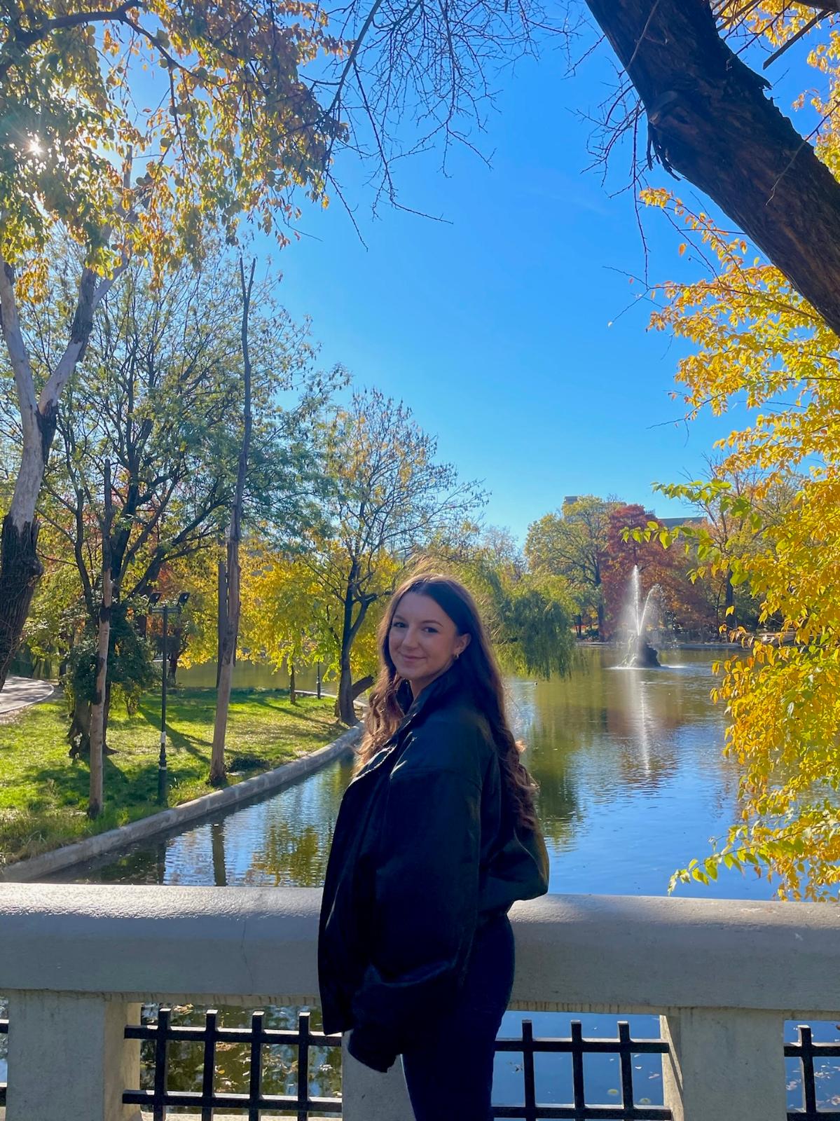 Brooke stands side ways and in the background is a park and a river with a fountain