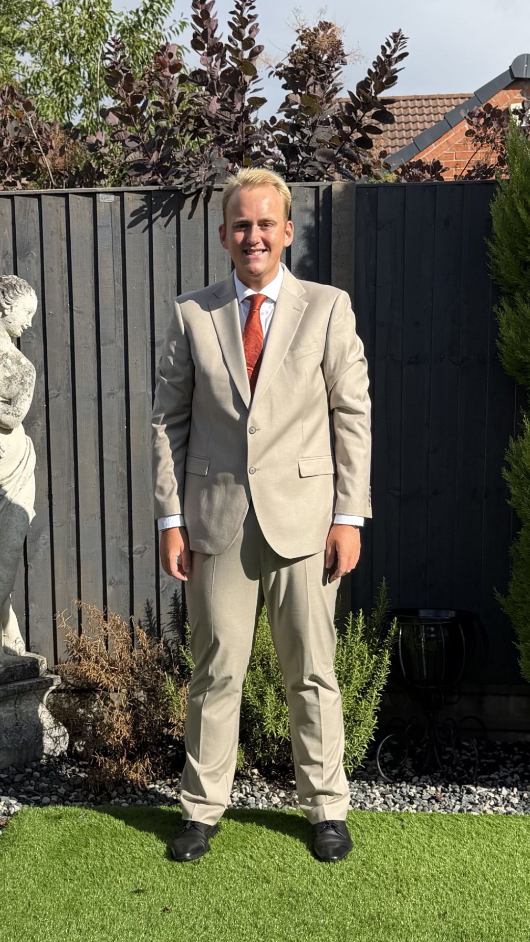 Leon stands in a garden wearing a beige suit and red tie