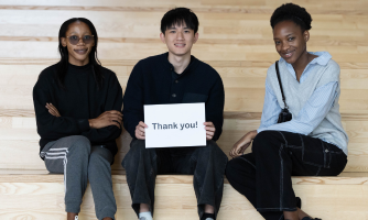 Three students sit on steps with one holding a piece of paper that reads Thank You
