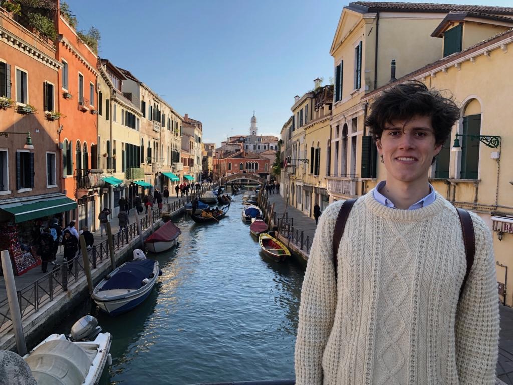 Matthew stands in a white jumper with a background of the Venice waterways