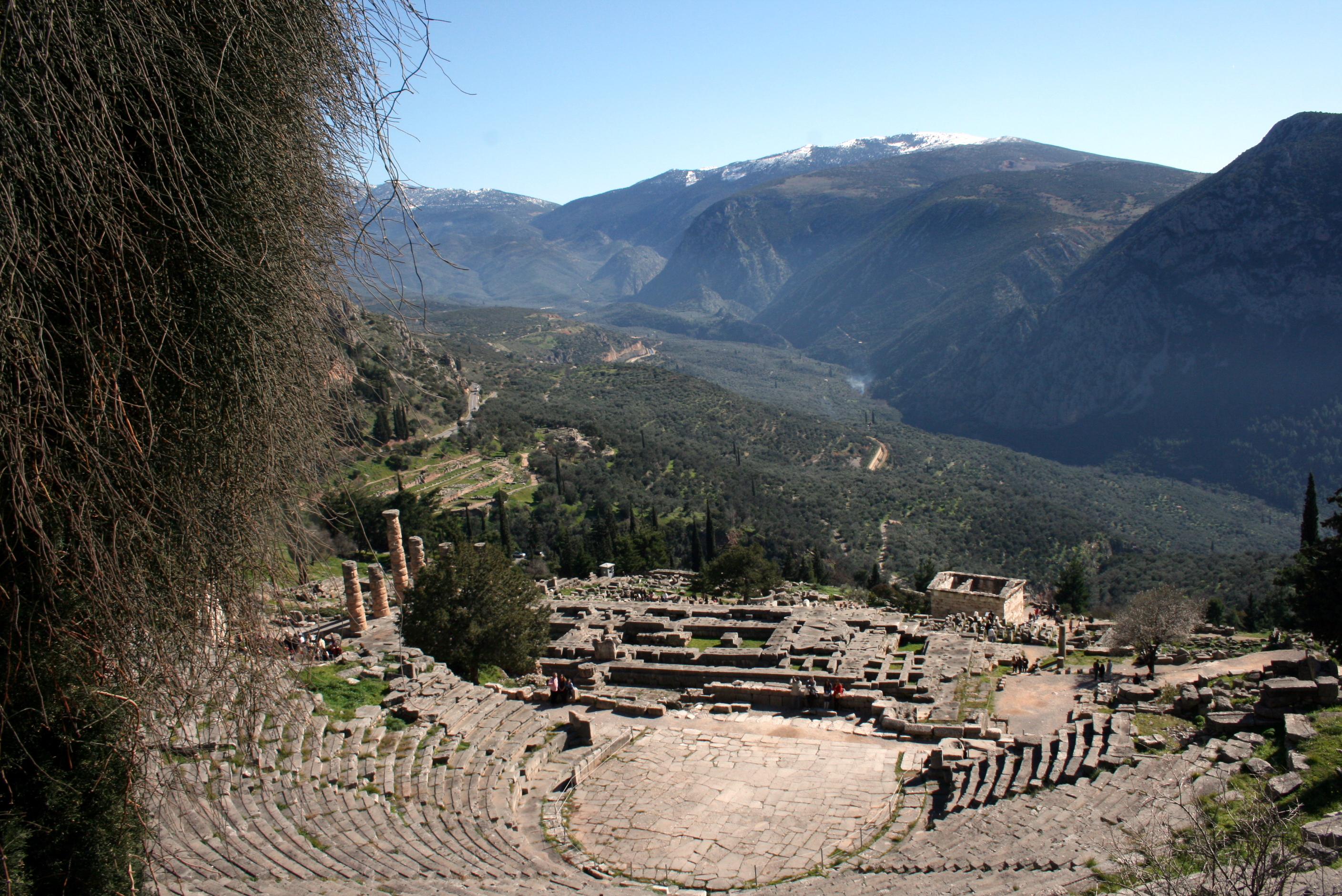 Photo of the Greek sanctuary at Delphi. Photo shows the ancient theater in the foreground, the temple of Apollo in the mid-ground, and the valley and mountains that surround Delphi. Photo by Conor Trainor