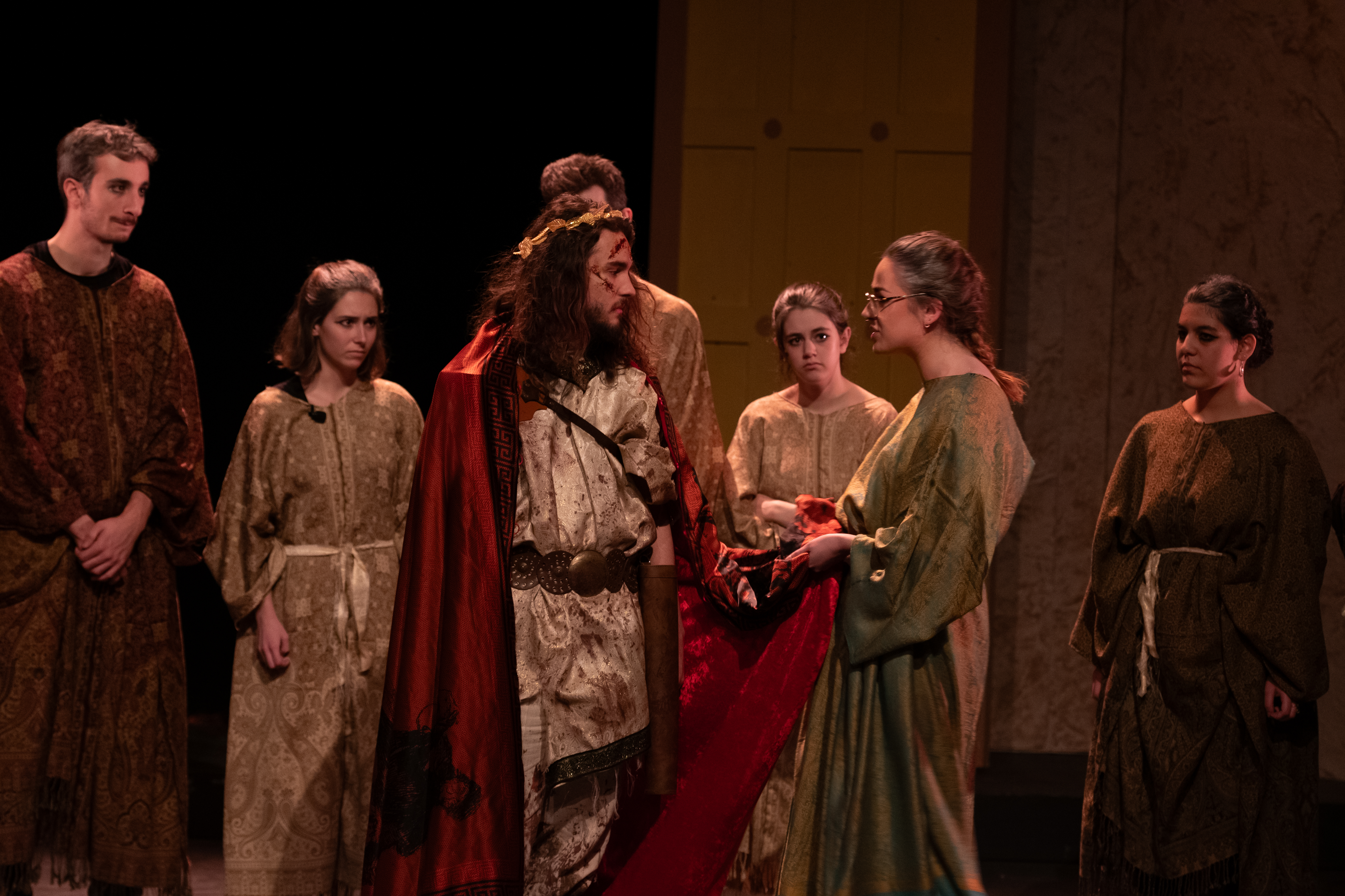 The chorus interrogating King Xerxes, reading from the scenes of defeat on his cape.