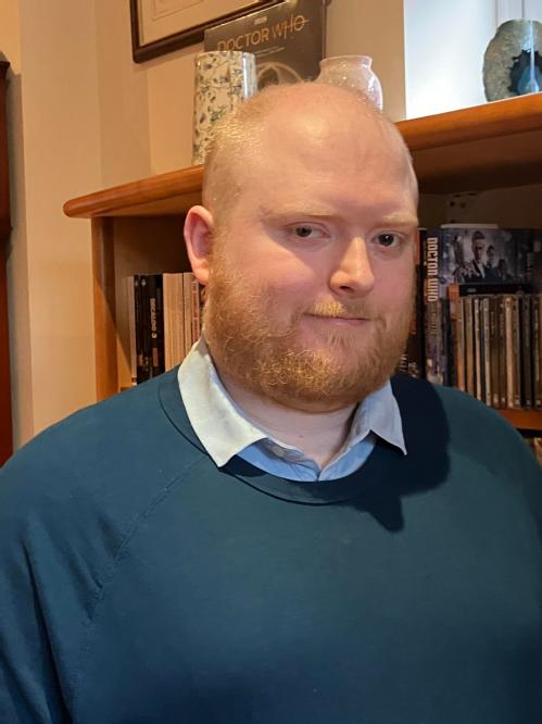 This image depicts Dr. Julian Richards, a white man in his thirties with a red beard and close cropped red hair. He is wearing a turquise jumper over a blue polo shirt, and is stood in front of a bookshelf.