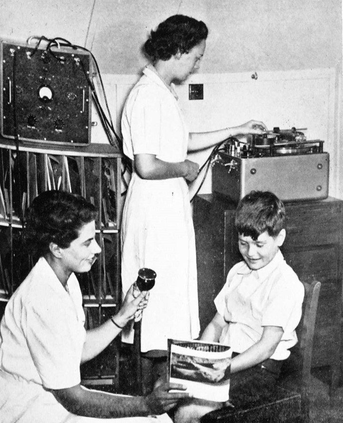 'The recording room'. One speech therapist holds a microphone to a boy reading a magazine, while another operates the magnetic reel-to-reel tape (1950s).