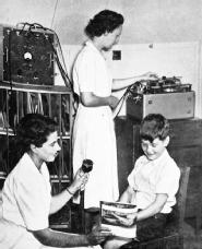 'The recording room'. One speech therapist holds a microphone to a boy reading a magazine, while another operates the magnetic reel-to-reel tape (1950s).
