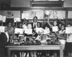 Black and white photo showing smiling young students of multiple ethnicities surrounded by books and other historical paraphernalia.