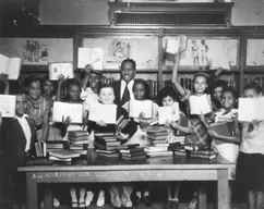 Black and white photo showing smiling young students of multiple ethnicities surrounded by books and other historical paraphernalia.