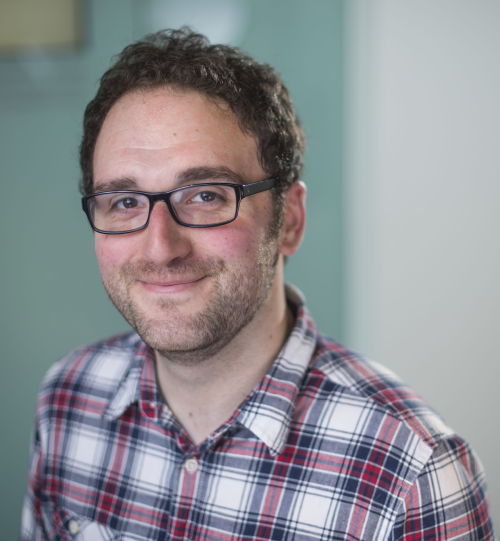Dr Simon Peplow is wearing a checked shirt and glasses and smiling at the camera