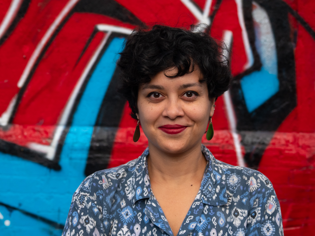 Profile photo of South Asian person with short black hair, green earrings, red lipstick and a blue patterned shirt with an open collar, standing in front of a wall with graffiti in bright red, blue, black and white