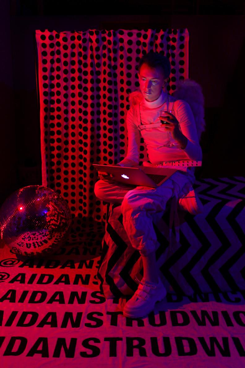 monochromatic pink photo of an androgynous subject wearing angel wings sat looking at a laptop, there is a disco ball and a curtain surrounding them and the floor is covered in writing that says @aidanstrudwick