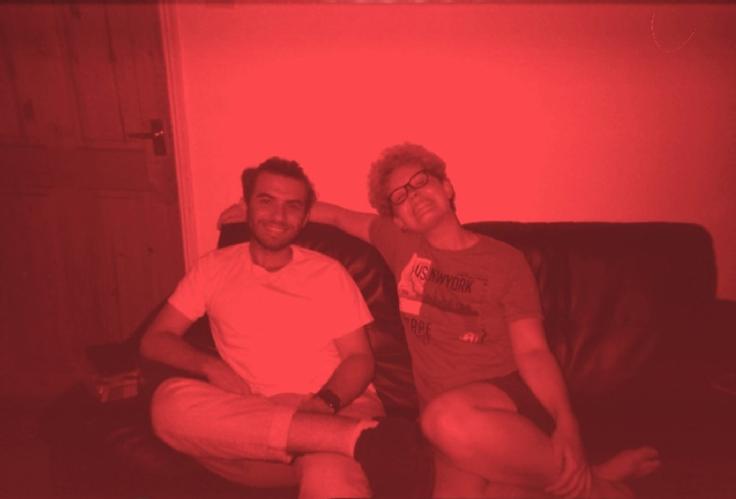 Amber's trans boyfriend, Oliver, (right) and their mutual friend, Mustafa, (left.) A red tinged photograph of the two arm in arm.