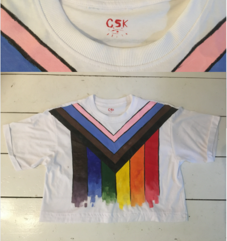 This image shows a tshirt that has been painted with the progress pride flag. The rainbow goes downwards with red on the right most wide and around the colour are the trans pride colours and the black and brown striped for POC inclusion. A second close up image shows the logo printed into the back collar of a C S K, the initials of the artist with an eyeball underneath