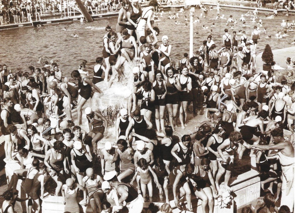 Crowds at Enfield Lido, 1934. Black and white photograph depicts crowds of people around an open-air swimming pool. Some sunbathers/swimmers are oblivious to the camera, while others notice it and stare back.