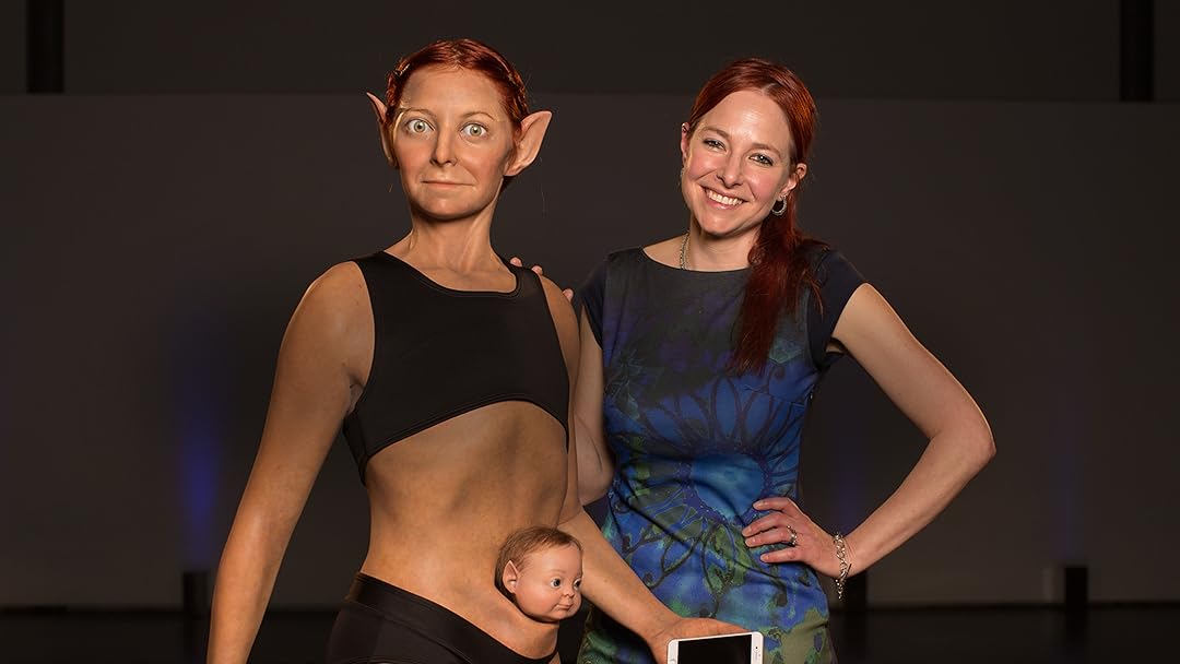 Alice Roberts, the paleopathologist and TV presenter, stands smiling next to a live-sized image of her as perfected by science. The perfect Alice has larger eyes, large and pointed ears, and a prominent abdominal pouch from which an infant's head (also with pointed ears) protrudes.