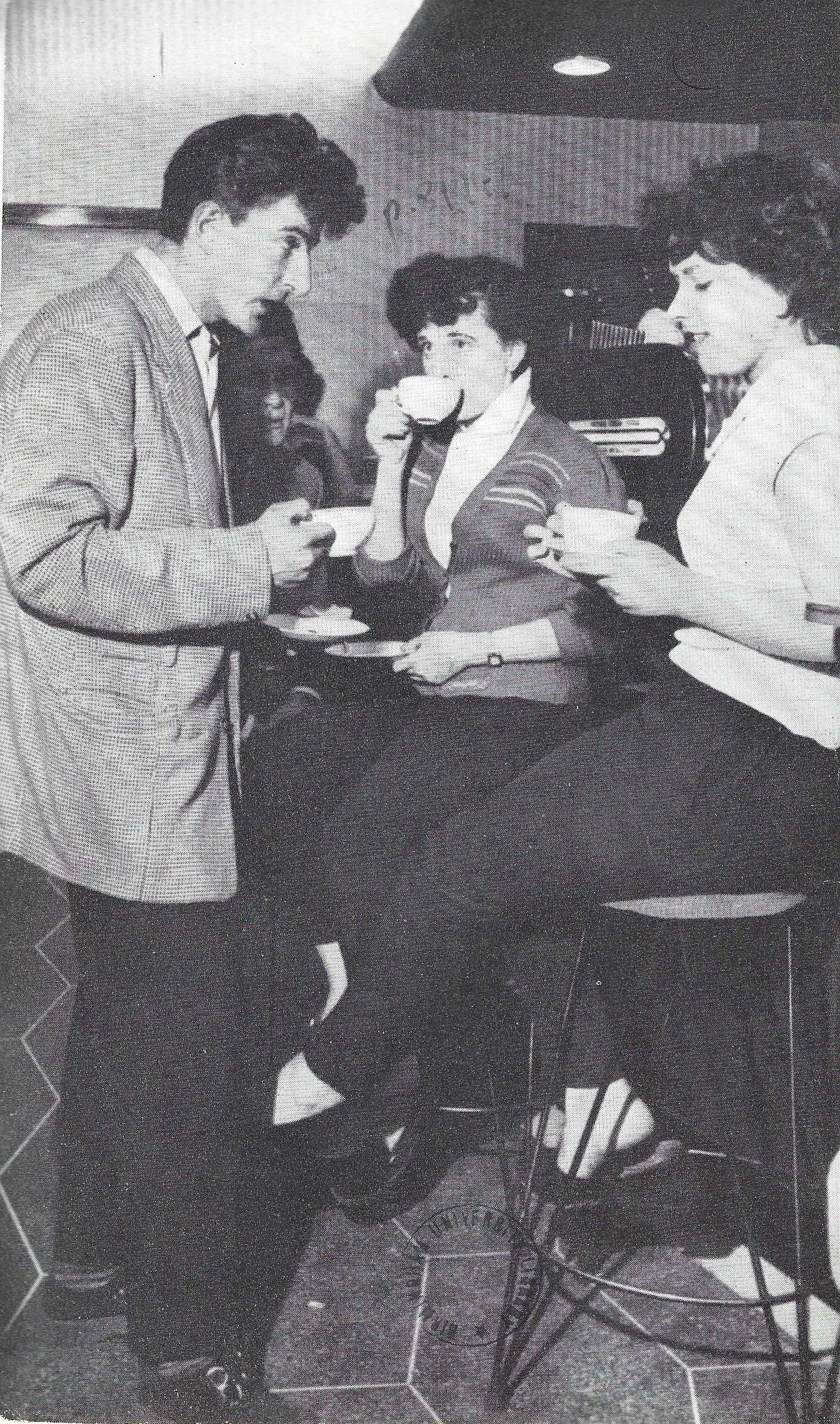 Black and white photograph depicts three young people in a coffee bar. Two young women with curly hair and wearing jumpers are sat on bar stools, obscuring the view of the bar itself. A young man in a light coloured jacket with styled hair stands facing them. All of them are holding, or sipping from, porcelain cups with saucers.