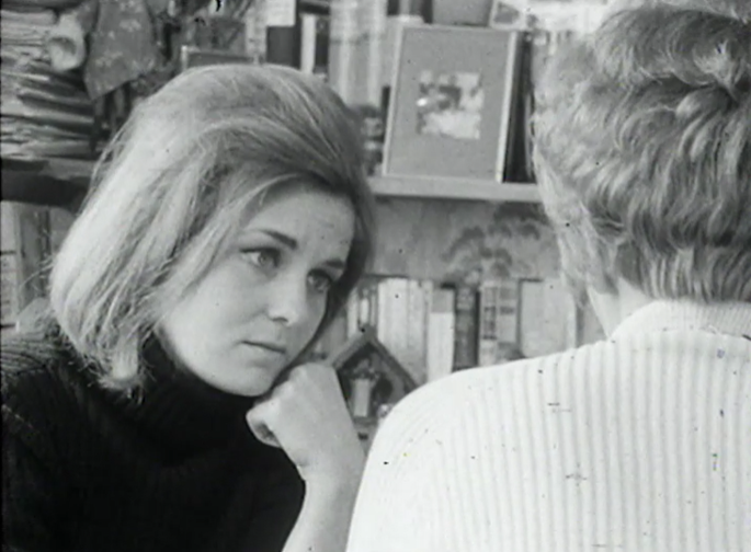 Black and white still. A close up of Angela Huth (left) a young woman with medium-length light-coloured hair, looking pensively and resting her head on her left hand. Her interviewee is on the right, with her back to the camera. She has short, wavy hair and a light-coloured jumper. The interview takes place in the interviewee's living room and the background features a set of display shelves with books and indistinct family ornaments and photographs.