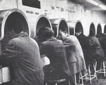 Black and white photograph shows a row of young people sat on bar stools along a wall with their heads underneath futuristic-looking metallic hoods. Above these is a sign that reads 'Melody Bar'.