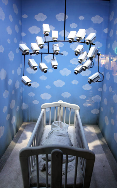 A baby's cradle sits beneath a mobile of CCTV cameras, against a background of blue summer skies. The image is a piece of art by Banksy, exhibited in his Croydon pop-up shop GDP in 2019