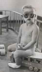 Black and white photograph shows a young infant sat on top of a toy bus, surrounded by railings (a sort of play pen) and other toys. He is wearing tinted goggles.