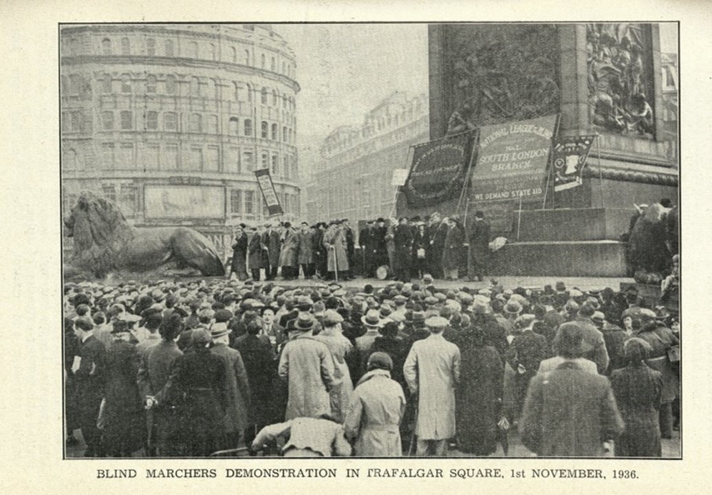 Black and white photograph: Nelson's column base is visible in the background, with one of the lion sculptures visible to the left. A large crowd of people in coats, hats and caps (mostly male with a few women) are looking towards the line of speakers at the base of the column.  Behind them are a few banners, with texts which are illegible at this distance.