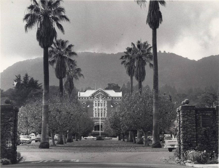 Black and white photograph of the home: an imposing, cuboid building viewed at the end of a drive lined by palm trees and cars. Mountains and clouds are visible in the background.