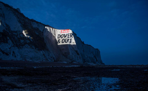 The image depicts a video projection onto a cliff face at Dover, which reads "The Sun. DOVER & OUT"