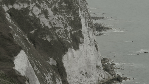 A view looking down at the white, chalk cliffs of Dover. The sea laps the bottom of the cliffs. The colours are muted and greyish.