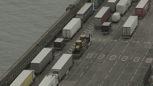 View down into the detail of a port area. Columns of trucks move in queues, diagonally through the frame. To the left a murky sea can be seen.