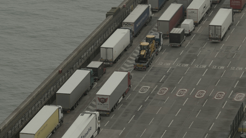 View down into the detail of a port area. Columns of trucks move in queues, diagonally through the frame. To the left a murky sea can be seen.