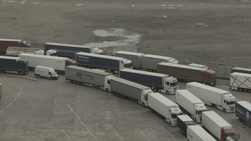 A view into a grey tarmac area through which columns of queuing trucks arc. The colours are muted and greyish.